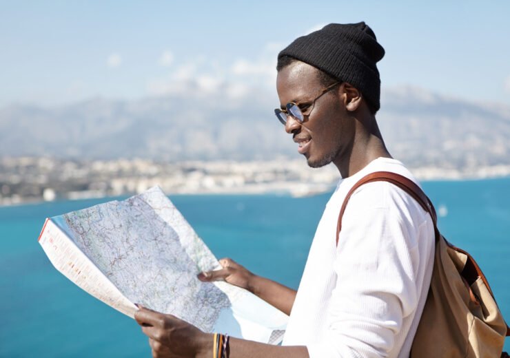 Side view of smiling trendy looking young dark-skinned hitchhiker studying paper map in his hands, planning route and stops during vacations abroad, vast azure sea and resort town in background